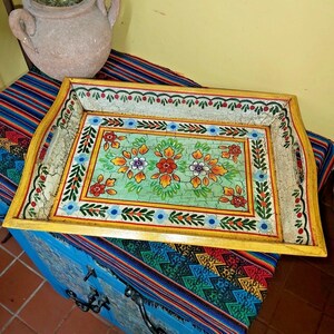 May include: A rectangular wooden serving tray with a yellow border and a central light green panel featuring hand-painted floral motifs in red, orange, and purple. The surface has a distressed crackle finish with decorative blue and green borders.