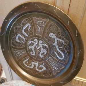 May include: A round brass tray with an intricate design featuring Arabic calligraphy in silver. The tray has a raised rim and a smooth surface.