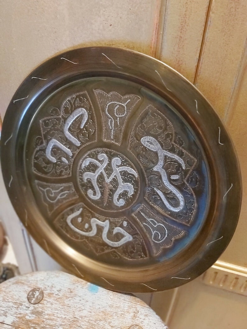May include: A round brass tray with an intricate design featuring Arabic calligraphy in silver. The tray has a raised rim and a smooth surface.