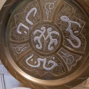 May include: A round brass tray with intricate silver-colored designs and Arabic calligraphy. The tray has a raised rim and a slightly textured surface.
