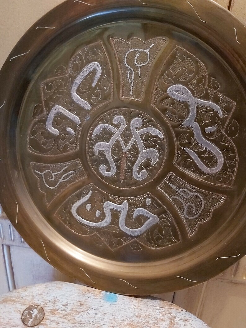 May include: A round brass tray with intricate silver-colored designs and Arabic calligraphy. The tray has a raised rim and a slightly textured surface.