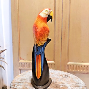 May include: A carved wooden parrot sculpture. The parrot has orange, red, yellow, and blue plumage. It is perched on a black stand. The sculpture is displayed on a distressed, round, light blue table.