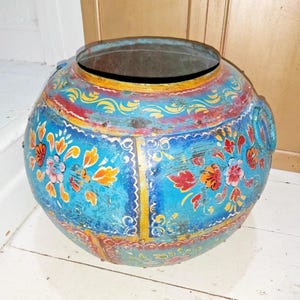 May include: A round, blue metal container with a wide opening and two side handles. It features floral patterns in red, orange, and yellow. The base and rim are accented with yellow and red trim.