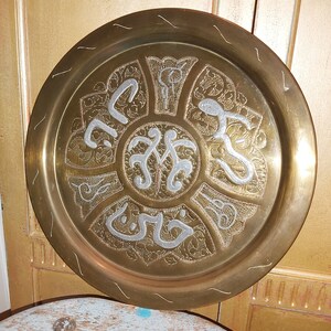 May include: A round brass tray with an intricate design of silver-colored Arabic calligraphy and floral patterns.