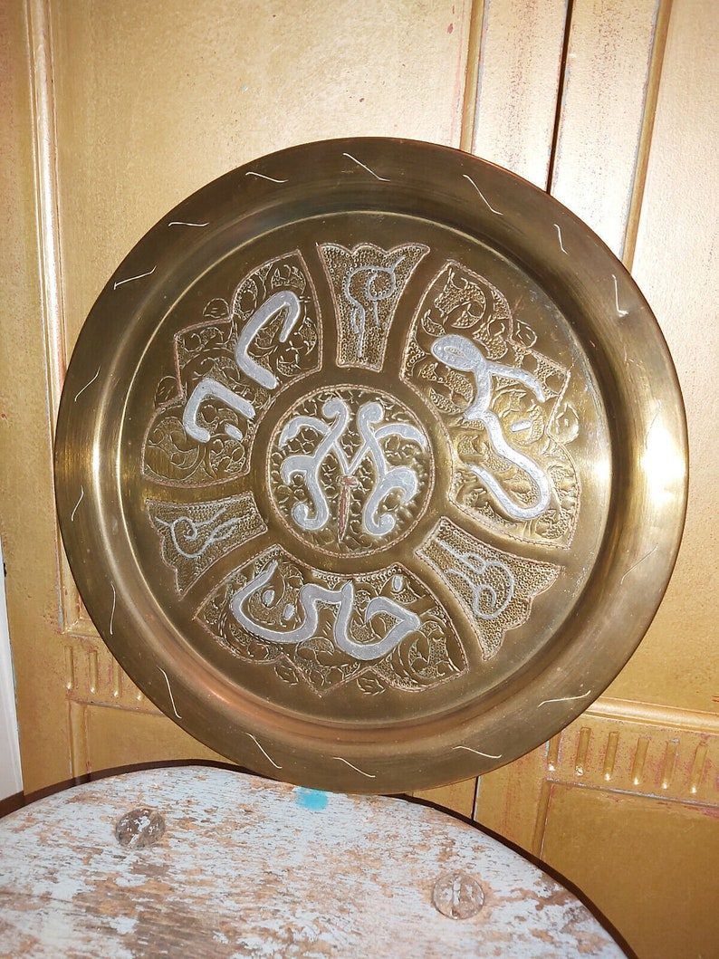May include: A round brass tray with an intricate design of silver-colored Arabic calligraphy and floral patterns.