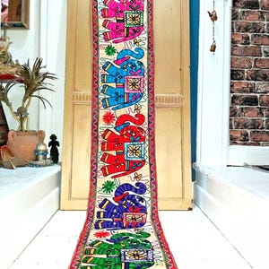 May include: A long, cream-coloured tapestry with a colourful embroidered design of five elephants. The elephants are in different colours, including pink, blue, red, purple, and green. The tapestry has a wavy border in red, white, and green.