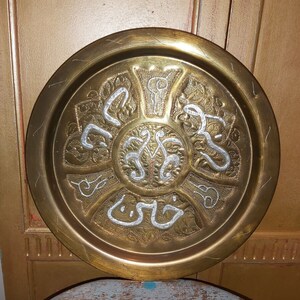 May include: A round brass tray with intricate silver filigree designs and Arabic calligraphy. The tray has a raised rim and a smooth, polished surface.