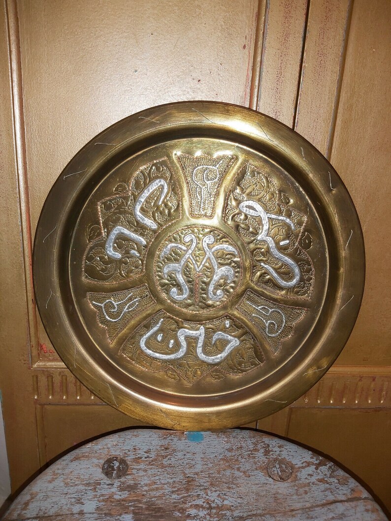 May include: A round brass tray with intricate silver filigree designs and Arabic calligraphy. The tray has a raised rim and a smooth, polished surface.