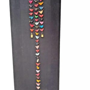 May include: A colourful string of fabric birds with bells at the bottom. The birds are in various colours, including red, blue, green, yellow, and pink. The string is hanging from a black background.