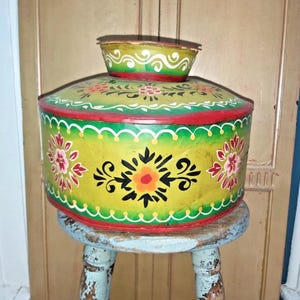 May include: A round, yellow tin container with a lid, decorated with red, green, and white floral patterns. The container sits atop a small, distressed blue stool. The lid has a smaller, matching container on top.