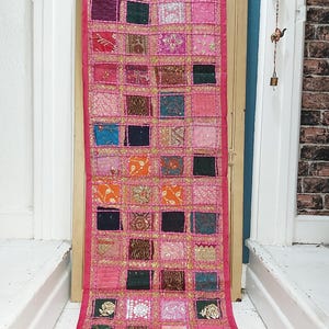 May include: A colourful pink wall hanging with a patchwork design. The rectangular textile features numerous squares in various colours, including pink, orange, green, and blue, each with unique patterns and embellishments. Gold trim outlines each square.