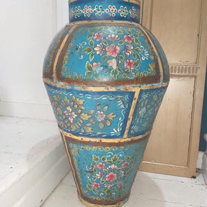 May include: A large, ornate, blue decorative vase with floral designs. The vase has a bulbous shape with a lid and a flared base. The floral patterns include pink, white, and yellow flowers. The vase is made of metal and has a distressed finish.