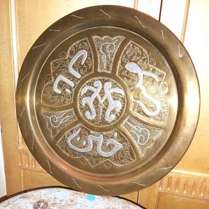 May include: A round brass serving tray with intricate engraved designs in silver. The center of the tray features a circular design with Arabic script.