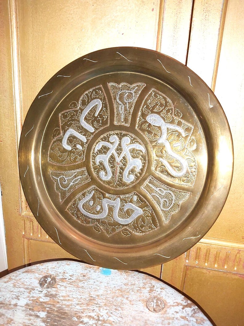 May include: A round brass serving tray with intricate engraved designs in silver. The center of the tray features a circular design with Arabic script.