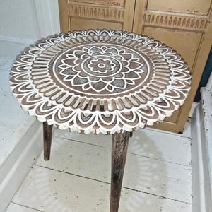 May include: A round wooden side table with a carved floral design on the tabletop. The table is painted in a brown and white colour scheme and has three slender legs. The table is approximately 50 cm in diameter.