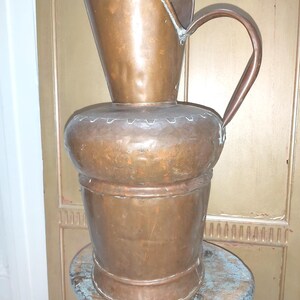 May include: An antique copper jug with a wide base and a tall, flared spout. The jug has a curved handle and sits on a small, distressed wooden stool. The copper has a warm, aged patina, suggesting its vintage. The jug is approximately 50 cm tall.