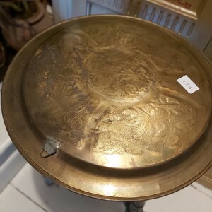May include: A round, gold-colored metal table top with an intricate, embossed design. The table top is attached to a wooden base.