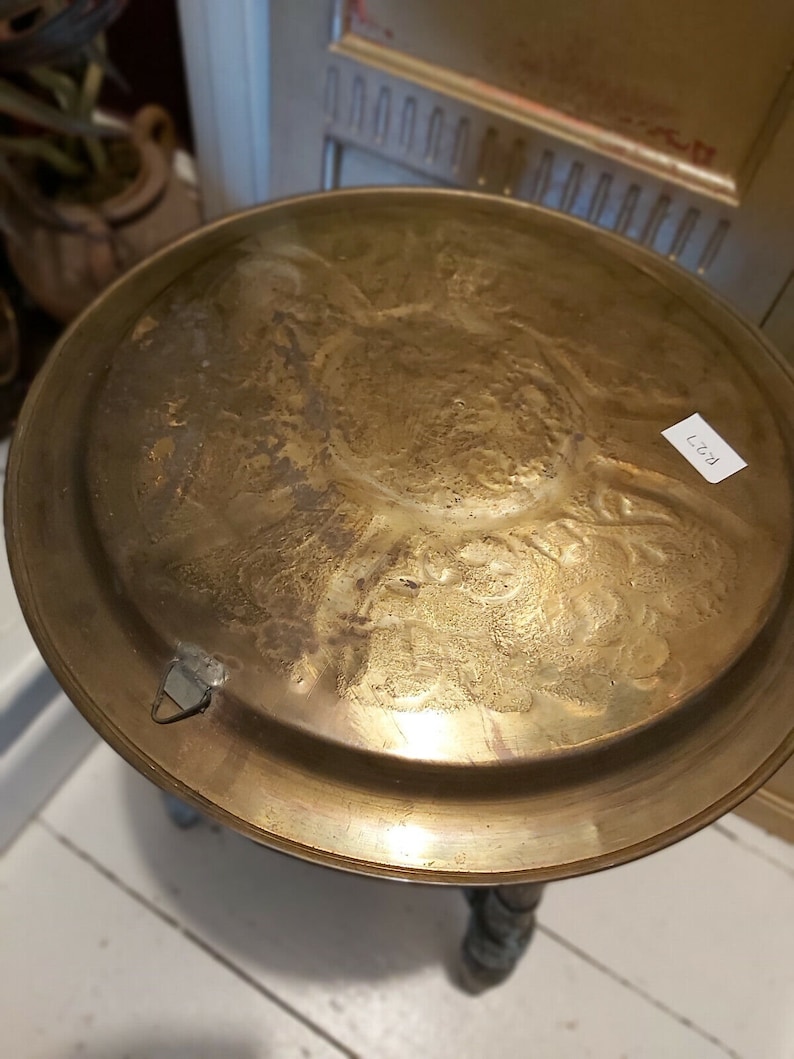May include: A round, gold-colored metal table top with an intricate, embossed design. The table top is attached to a wooden base.