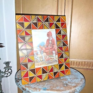 May include: A wooden picture frame with a geometric pattern of orange, red, brown, and yellow triangles. The frame is holding a picture of a man in a turban sitting on a rug with birds.