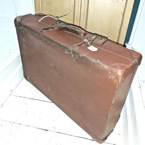 May include: A brown vintage suitcase with a leather handle and metal clasps. The rectangular case has reinforced corners and a slightly worn appearance, suggesting age and use. A small tag is attached to the clasp.