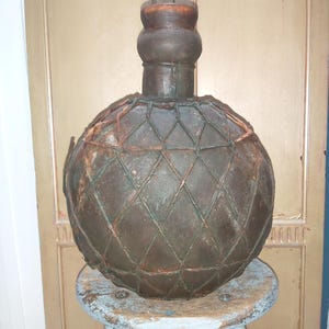 May include: A vintage, round, brown bottle with a textured surface and a diamond-patterned net overlay. The bottle has a cylindrical neck and a wooden stopper. It is displayed on a small, distressed, light blue stool.