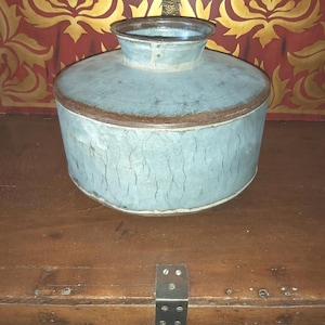 May include: A vintage, pale blue metal container with a cylindrical body and a flared top. The container has a dark brown rim and a distressed, crackled texture. It sits on a wooden surface.