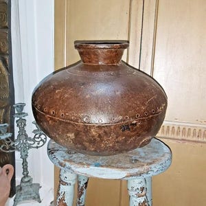 May include: A large, round, brown ceramic pot with a wide mouth and a slightly flared rim. The pot has a distressed, aged look with a textured surface. It is sitting on a light blue wooden stool with distressed paint.