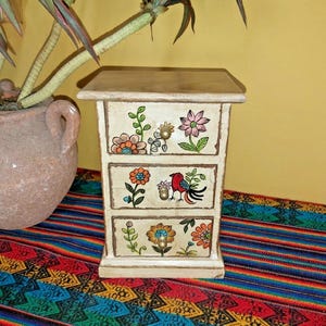 May include: A small, cream-coloured wooden chest of drawers with three drawers. Each drawer is decorated with colourful hand-painted floral designs and a bird. The chest sits on a vibrant, patterned textile, with a potted plant in the background.