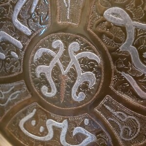 May include: A round, brass tray with intricate, silver-colored designs. The center of the tray features a stylized, silver-colored design with a small, brown nail in the center.