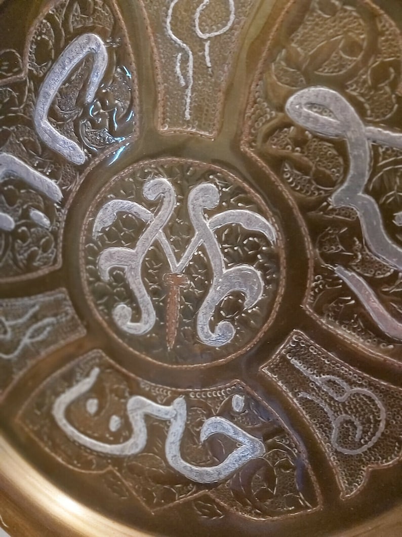 May include: A round, brass tray with intricate, silver-colored designs. The center of the tray features a stylized, silver-colored design with a small, brown nail in the center.