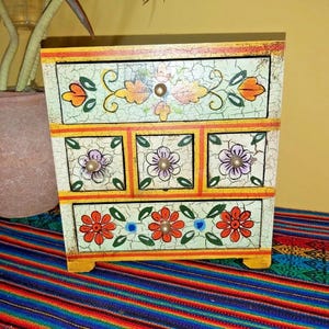 May include: A small wooden chest of drawers featuring a crackled off-white finish with hand-painted floral motifs in orange, purple, and green. The unit has one wide top drawer, three smaller middle drawers, and one wide bottom drawer with knobs.