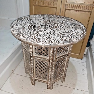 May include: A round, wooden side table with a white, carved floral design on the tabletop. The table has a brown base with intricate cut-out patterns and stands approximately 50 cm tall.