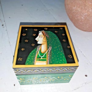 May include: A small wooden box featuring a painted design of a woman in a green sari. The box has a black background with gold accents and a green lid.