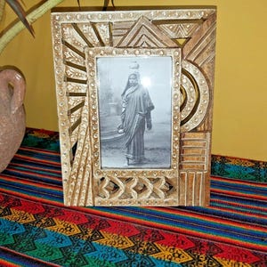 May include: A light brown wooden picture frame with a detailed geometric design. The frame contains a black and white photograph of a person. The frame rests on a colourful, patterned textile.