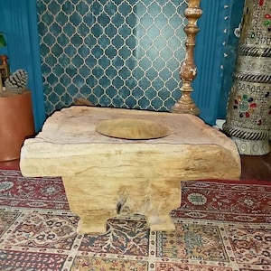 May include: A rustic wooden stool with a carved top and four legs. The stool is made of natural wood and has a distressed finish. It is sitting on a colourful rug.