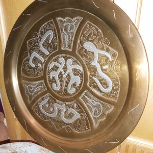May include: A round brass tray with intricate silver-colored designs and Arabic calligraphy. The tray has a raised rim and a smooth, polished surface.