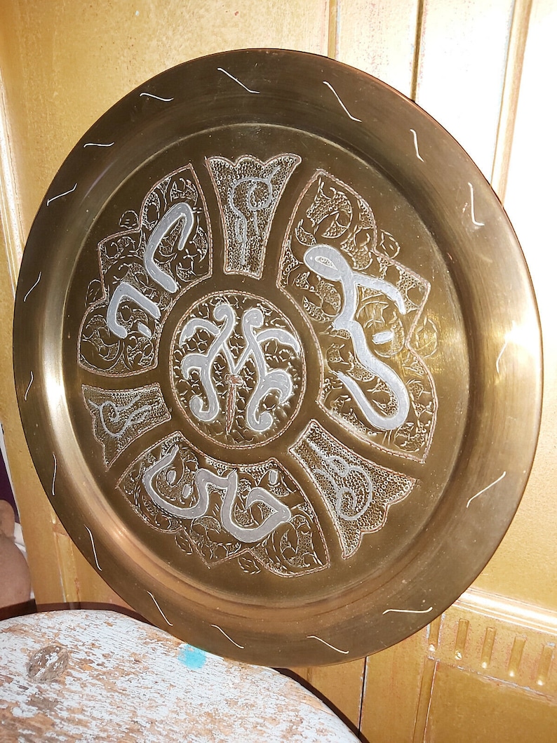 May include: A round brass tray with intricate silver-colored designs and Arabic calligraphy. The tray has a raised rim and a smooth, polished surface.