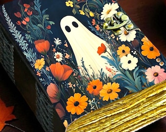 May include: A decorative book with a dark blue cover featuring a ghost illustration surrounded by colorful flowers. The book's pages have a gold-colored edge. A carved pumpkin and orange leaves are in the background, creating a Halloween theme.