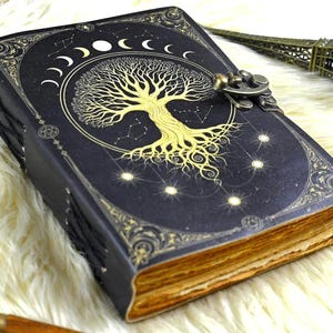 Puede incluir: Un diario de cuero negro con un diseño dorado de árbol de la vida, fases lunares y detalles de constelaciones. El diario tiene un cierre de metal y páginas envejecidas. La cubierta está decorada con detalles dorados.