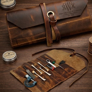 Peut inclure: Un étui à outils en cuir marron, ouvert pour révéler divers outils, dont une lime, des tournevis et une clé. L'étui est fermé par une lanière en cuir et un œillet en laiton. Le rabat supérieur est gravé d'initiales et du texte "MADE FOR ALBERT W. HARRISON CUSTOM ENGRAVED EST. 2024".