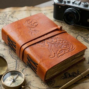 May include: A brown leather journal with an embossed wolf design on the cover. The journal is secured with a leather strap and features stitched binding. A vintage camera and compass are in the background.
