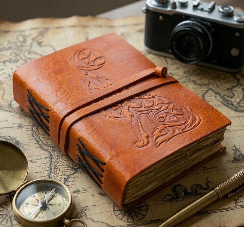 May include: A brown leather journal with an embossed wolf design on the cover. The journal is secured with a leather strap and features stitched binding. A vintage camera and compass are in the background.