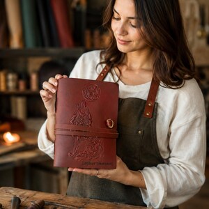 May include: A burgundy leather journal with a wolf and moon design, secured by a leather strap and metal clasp. The journal has the name "Zepher Swenson" embossed on the cover. The journal is held by a person wearing a white shirt and brown apron.