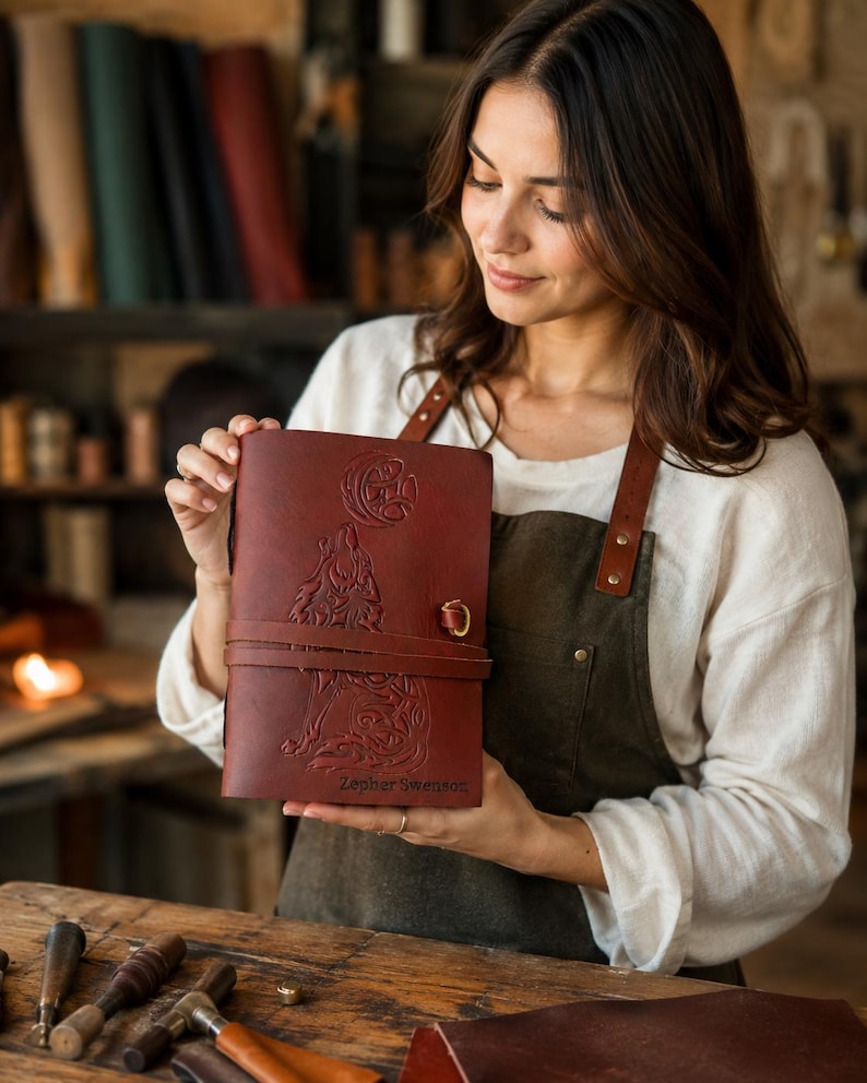 May include: A burgundy leather journal with a wolf and moon design, secured by a leather strap and metal clasp. The journal has the name "Zepher Swenson" embossed on the cover. The journal is held by a person wearing a white shirt and brown apron.