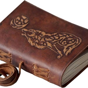 May include: A brown leather journal with a wolf howling at the moon design. The cover features an embossed wolf and moon, with stitched detailing and a leather tie closure. The journal is a rustic style.