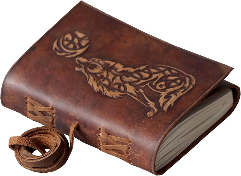 May include: A brown leather journal with a wolf howling at the moon design. The cover features an embossed wolf and moon, with stitched detailing and a leather tie closure. The journal is a rustic style.
