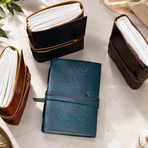 May include: An assortment of leather-bound journals in various colors, including brown, black, and teal. Each journal features a strap closure and contains a stack of white paper. Some journals have embossed designs on the cover.