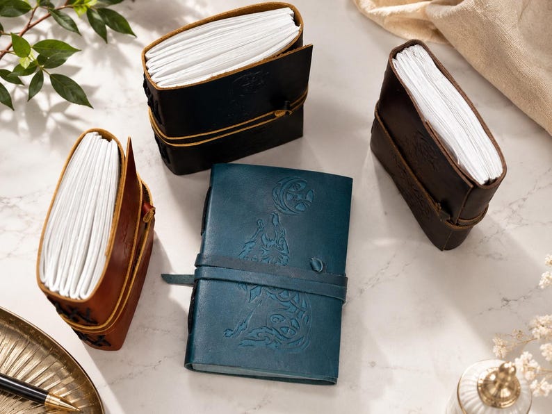 May include: An assortment of leather-bound journals in various colors, including brown, black, and teal. Each journal features a strap closure and contains a stack of white paper. Some journals have embossed designs on the cover.