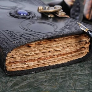 May include: A black leather journal with a decorative metal clasp and a blue gemstone. The journal has a worn, aged look and is open to reveal blank pages. A gold and black fountain pen is lying next to the journal.