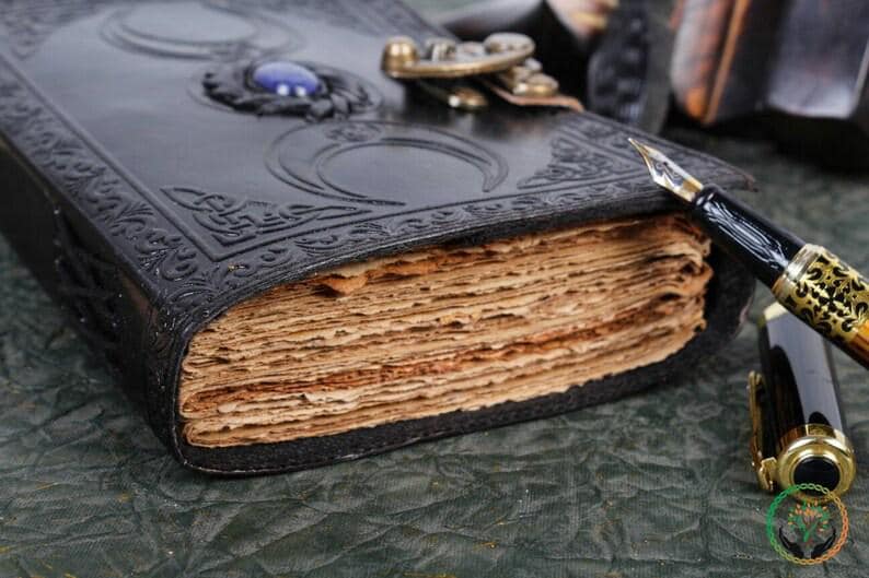 May include: A black leather journal with a decorative metal clasp and a blue gemstone. The journal has a worn, aged look and is open to reveal blank pages. A gold and black fountain pen is lying next to the journal.